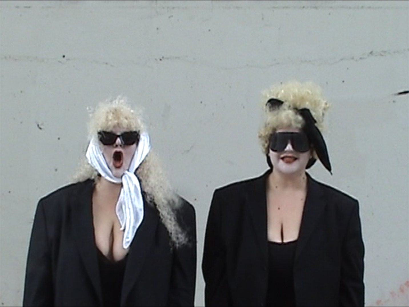 Two women with curly blonde hair, wearing black sunglasses and black outfits, standing against a plain wall. One woman on the left has her mouth open wide, and the other has her tongue out slightly. Both are wearing headbands or scarves and have exag