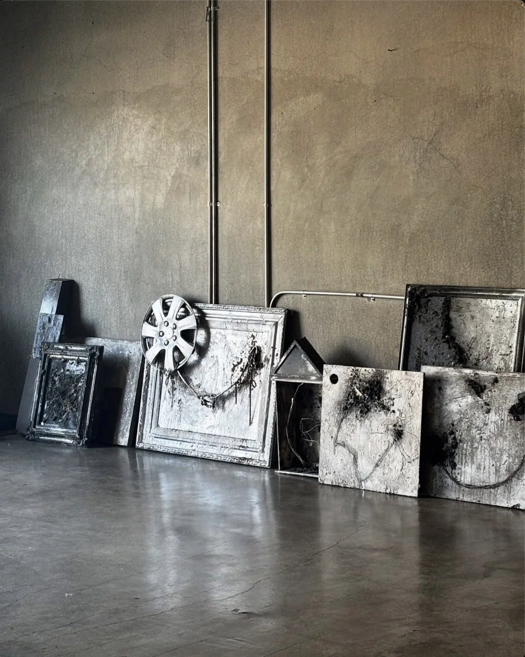A collection of blackened, charred picture frames and mirrors leaning against a beige wall in an empty room with a concrete floor. One frame has a visible wheel rim attached to it.