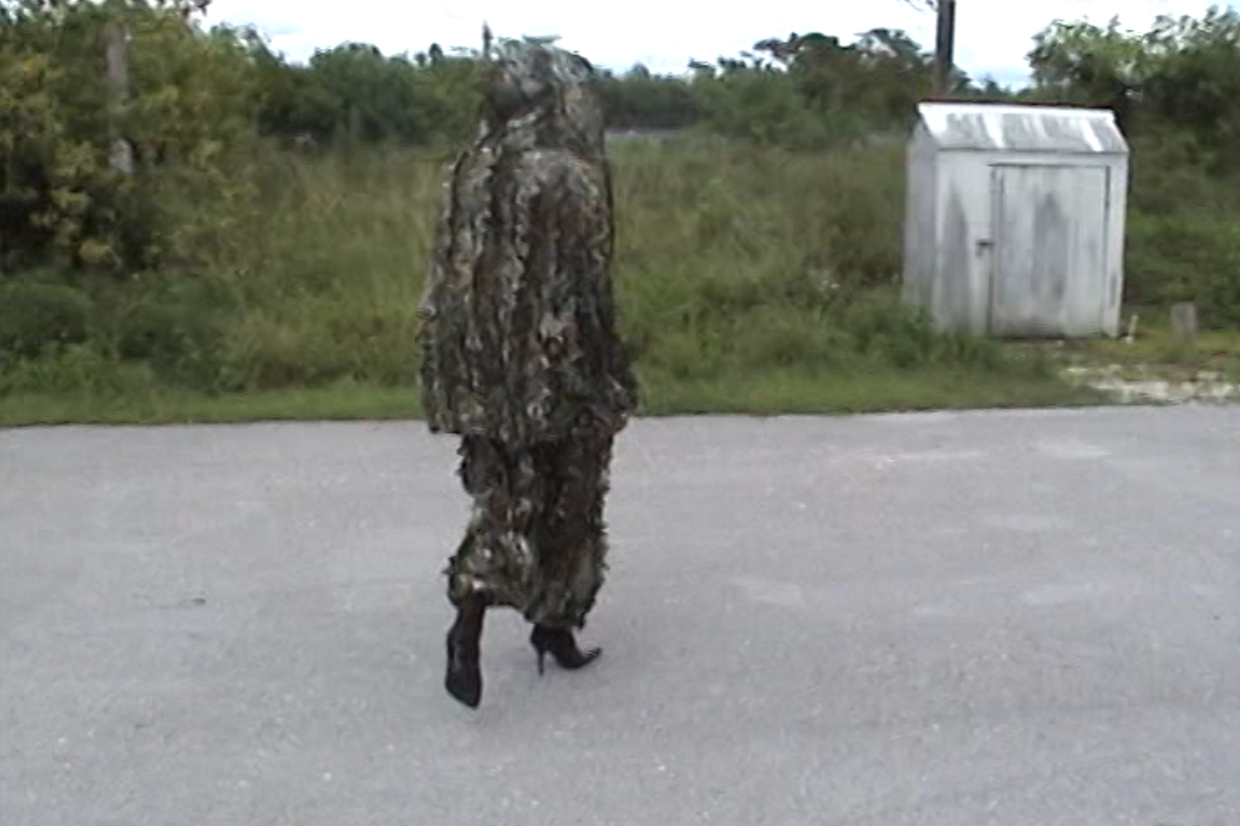 Person in camouflage clothing walking on the street with a grassy area and a small metal shed in the background.
