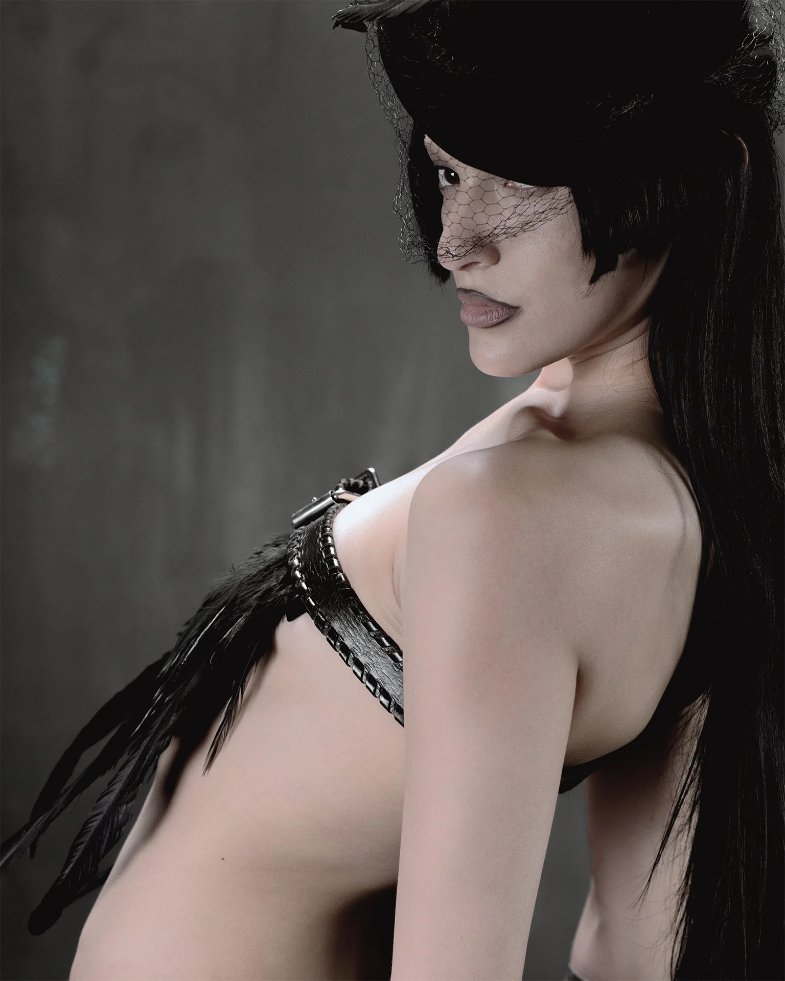 A woman with black hair, wearing a black lace veil and black feathered accessories, is posing sideways against a dark background.