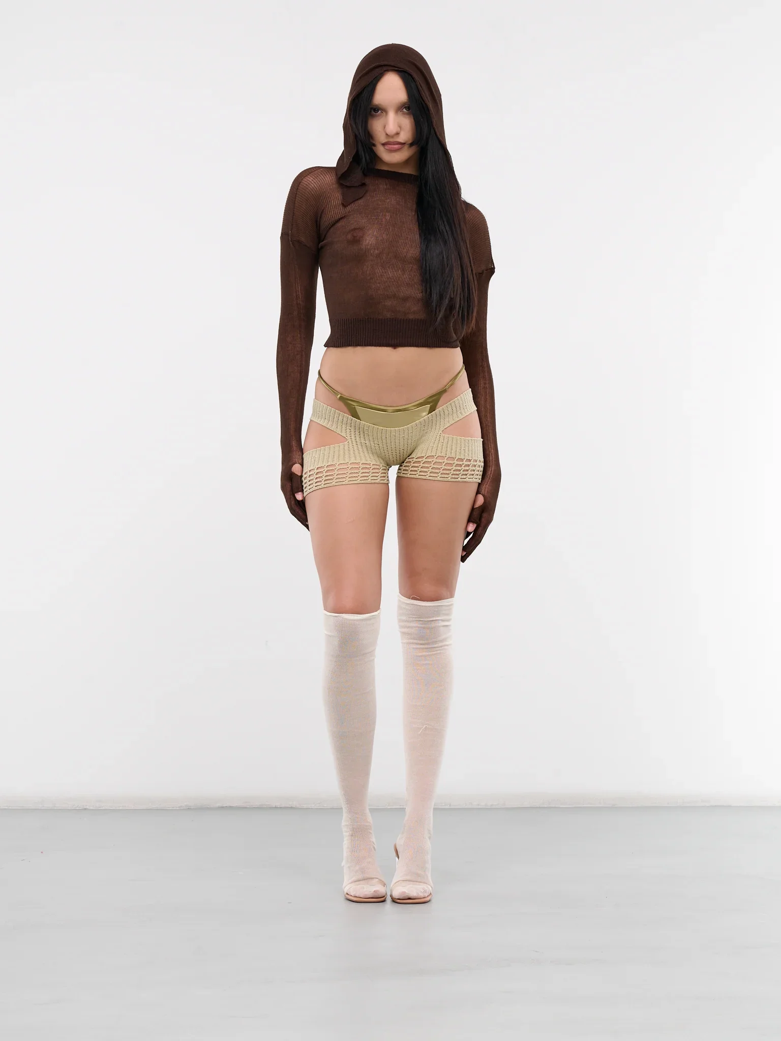 Woman standing against a plain white background, wearing a brown sheer hooded sweater, beige cut-out shorts with gold details, thigh-high beige socks, and high heels, with long dark hair.