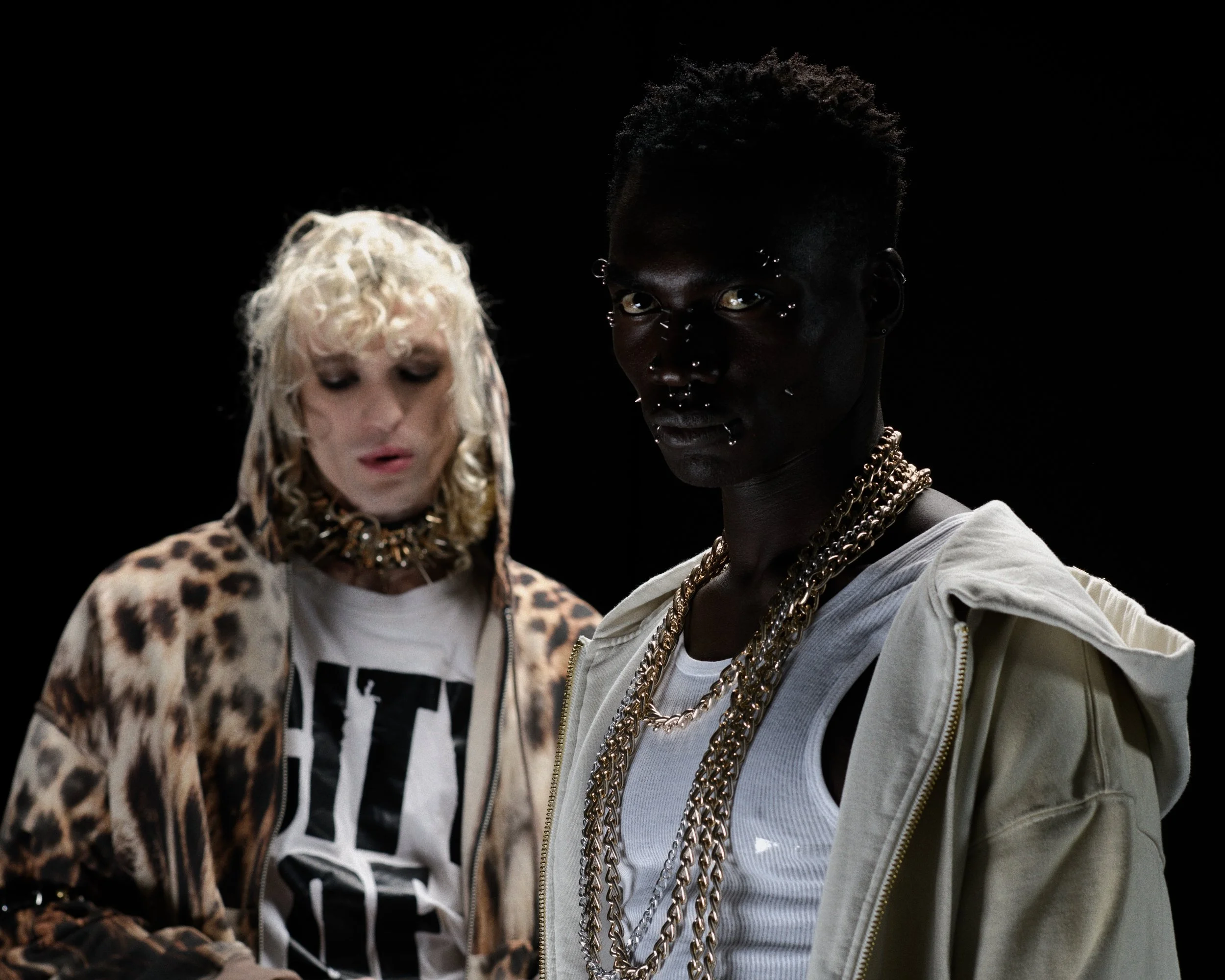Two individuals, one with blonde curly hair wearing a leopard-print jacket and graphic t-shirt, and the other with short dark hair and multiple chains, dressed in white and beige, standing against a black background.