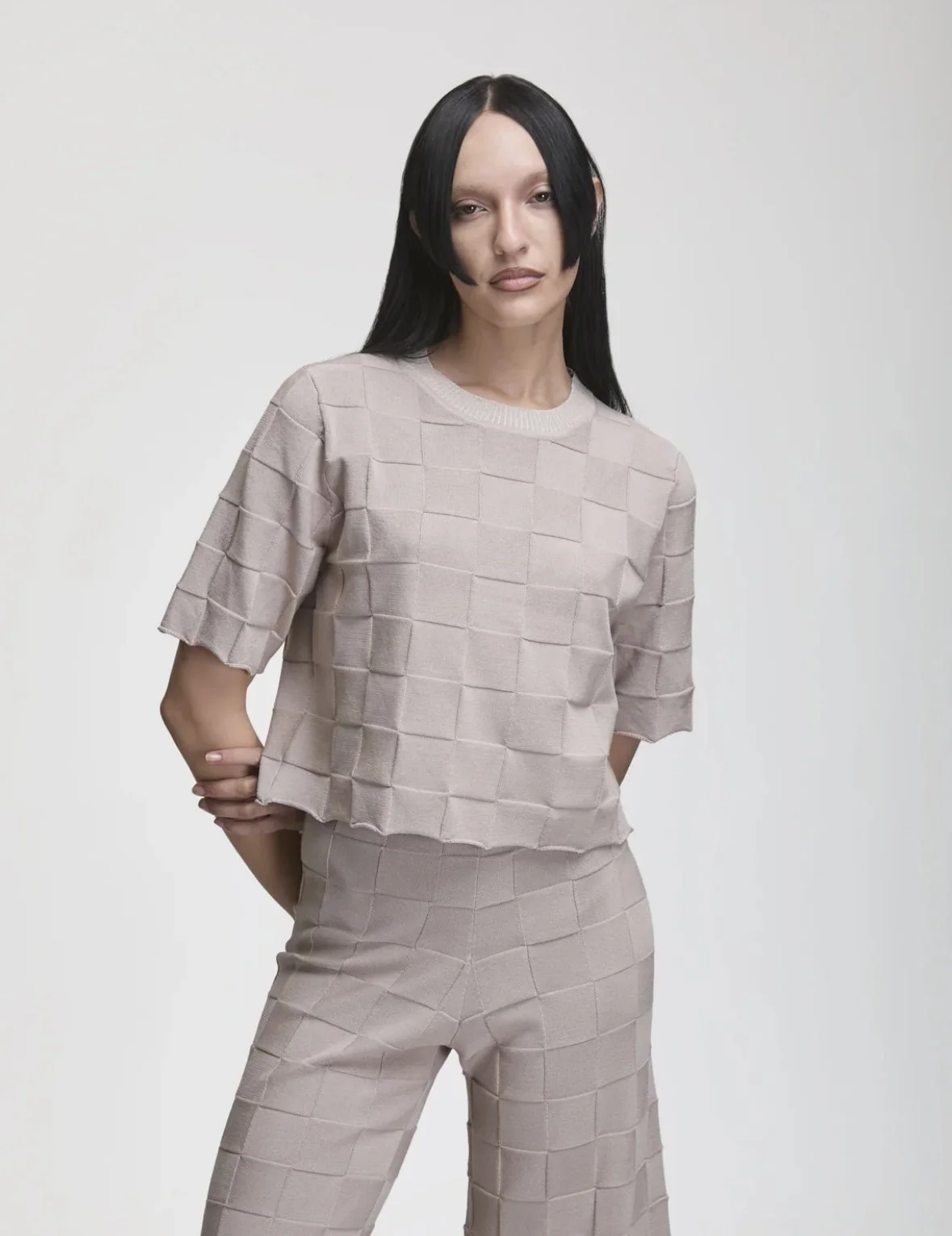 A woman with long black hair wearing a beige matching grid-patterned top and pants standing against a plain light background.