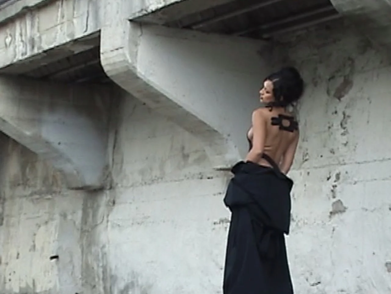 A woman with short black hair, topless, standing against a concrete wall, with her back turned to the camera, and black clothing gathered around her waist.