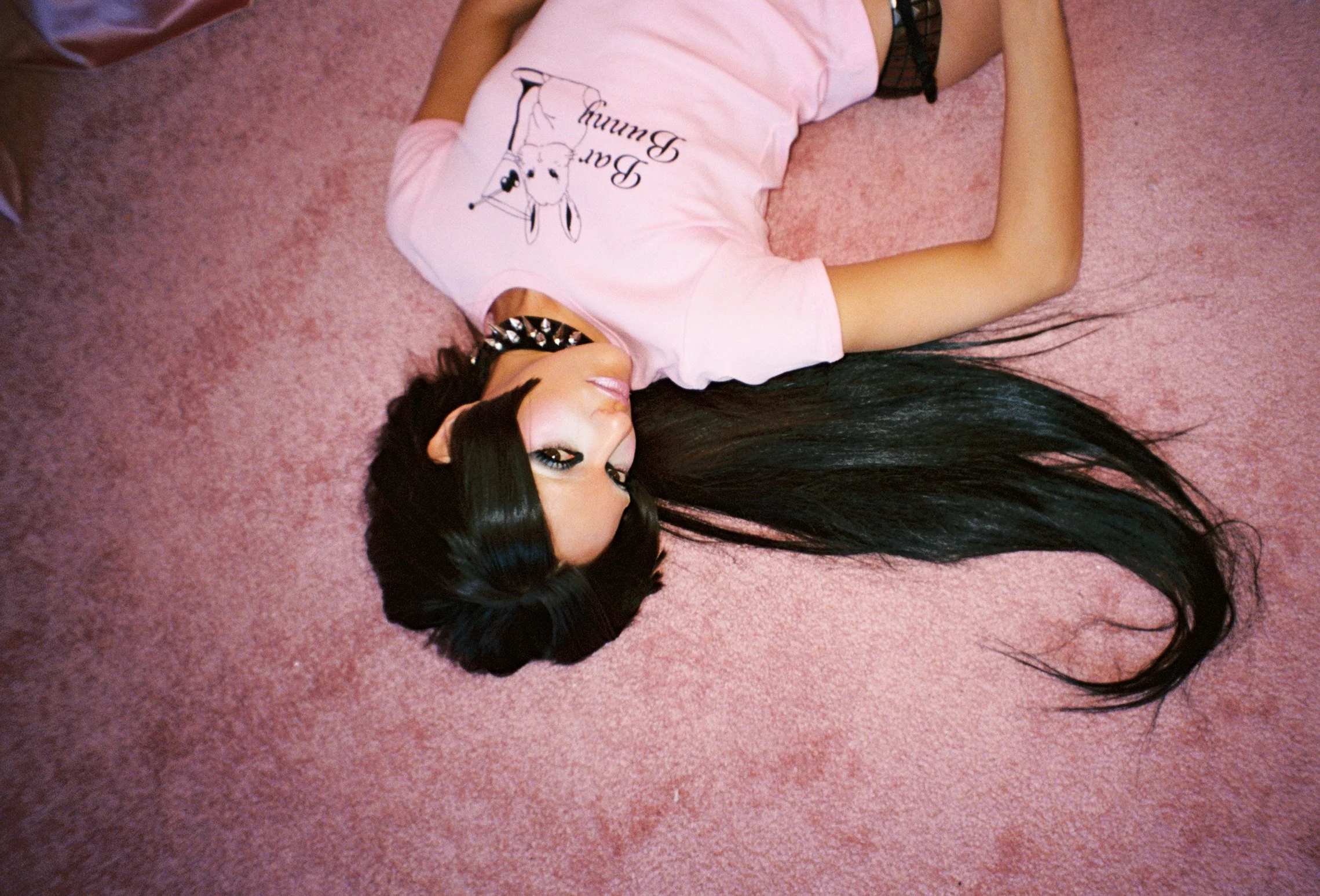 A woman with long black hair lying on a pink carpet, wearing a pink T-shirt with a bunny graphic and the words 'Bae Bunny,' and a black studded choker.