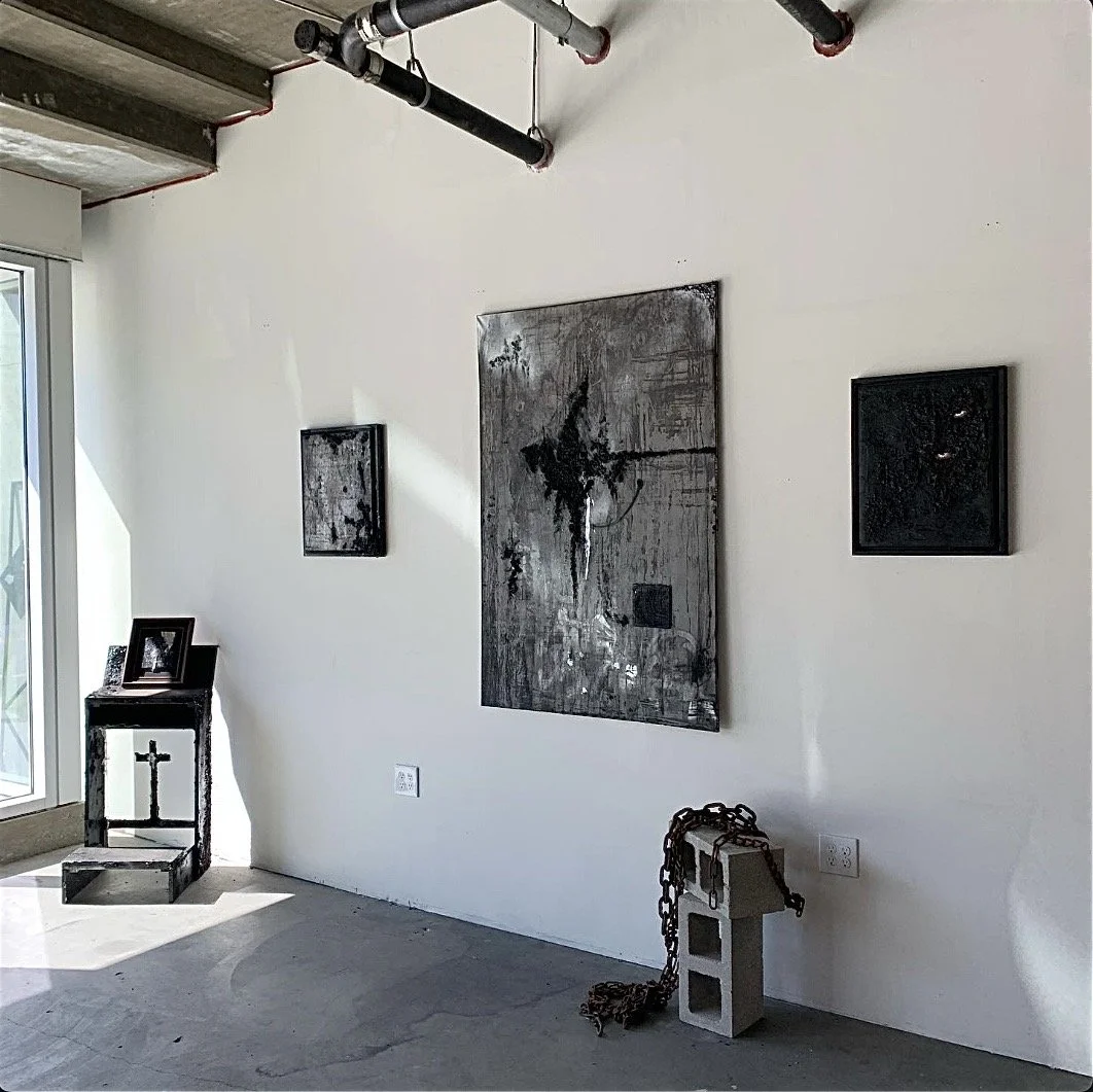 Modern interior with abstract artwork on white wall, black and white religious cross on black wooden stand, chain draped over cinder block on concrete floor, and art collection displayed near sliding glass door.