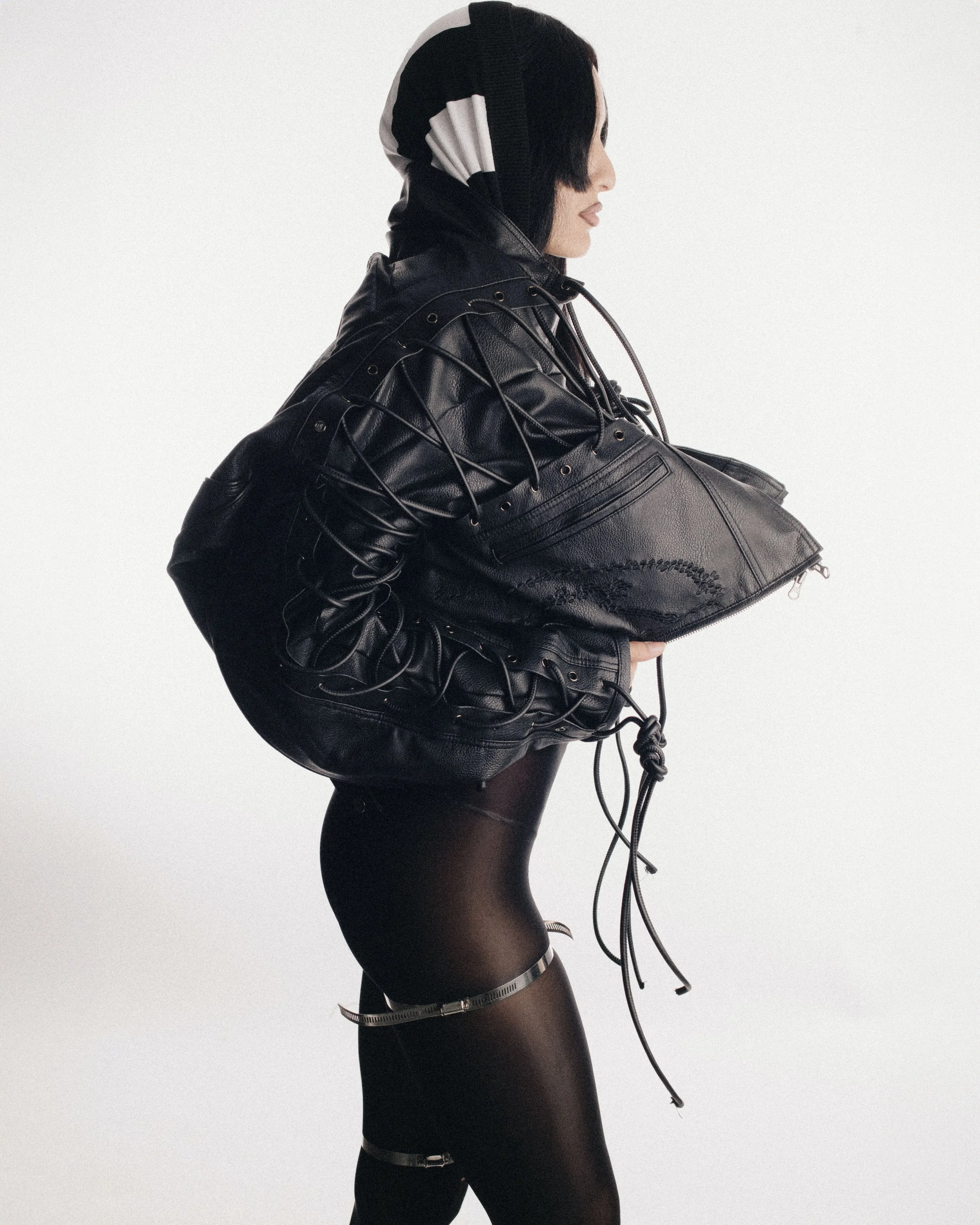 Person wearing a black leather jacket with laced details, holding a black handbag, in black tights with zipper details, and a striped hooded scarf, standing sideways against a plain white background.