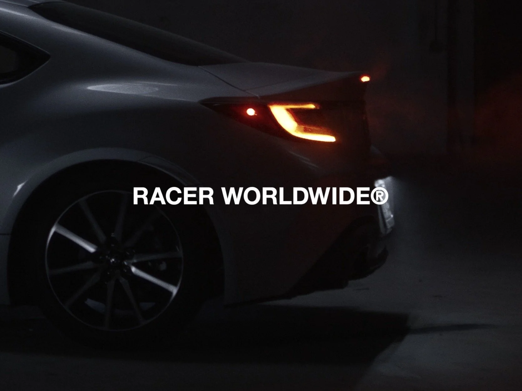 Rear side of a gray sports car with illuminated tail lights, parked in darkness, with the text 'RACER WORLDWIDE®' overlaid.