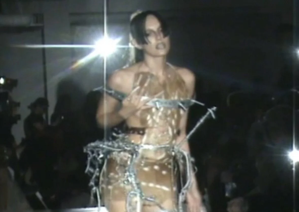 A woman with short black hair and glasses wearing a metallic outfit, holding a microphone, standing in front of a bright light and a dark background.