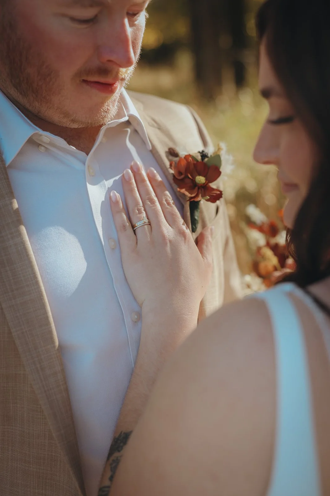A bride and groom are close together outdoors, with the bride's hand on the groom's chest showing her wedding ring, and they appear to be sharing an intimate moment on their wedding day.