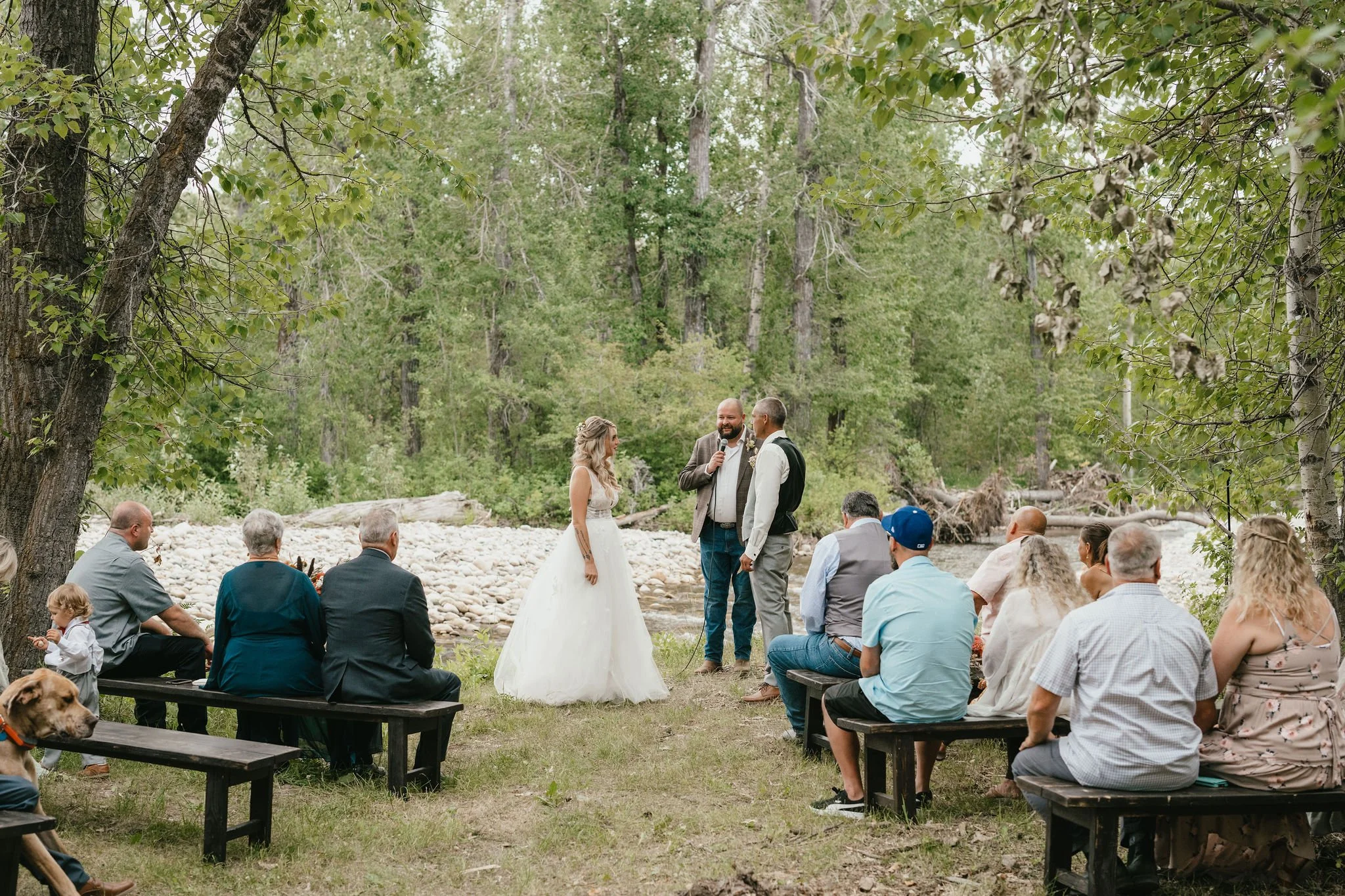 A wedding ceremony outdoors in a wooded area near a creek, with the bride and groom exchanging vows in front of friends and family.