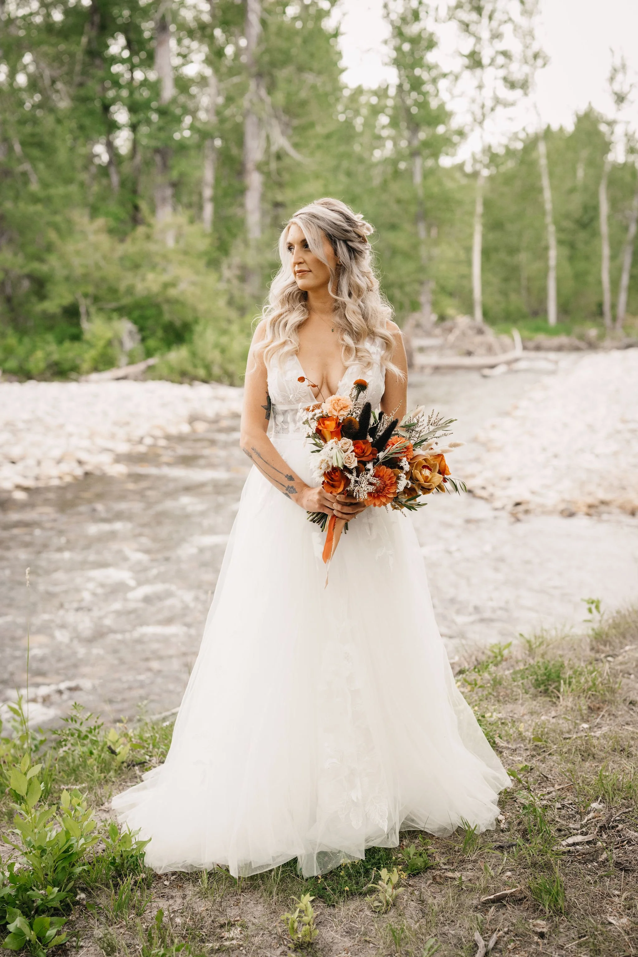A woman in a wedding dress holding a bouquet of flowers outdoors near a river with trees in the background.