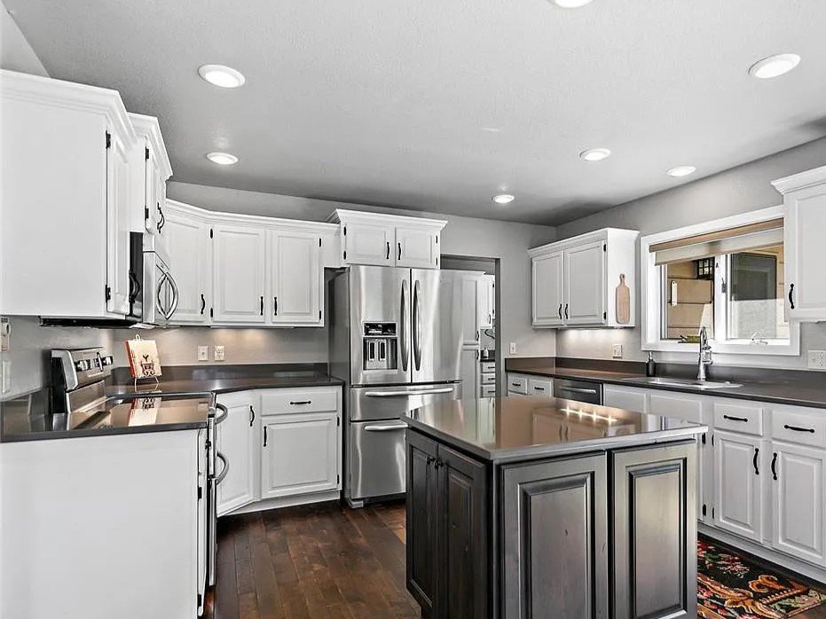 Modern kitchen with white cabinets, dark countertops, stainless steel refrigerator and oven, island in the center, window above sink, hardwood flooring, and recessed ceiling lights.