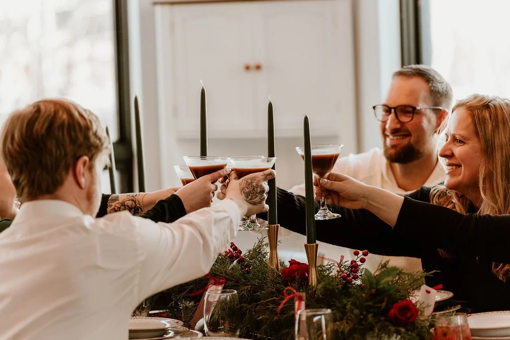 People raising glasses for a toast at a holiday dinner table decorated with greenery and red flowers.