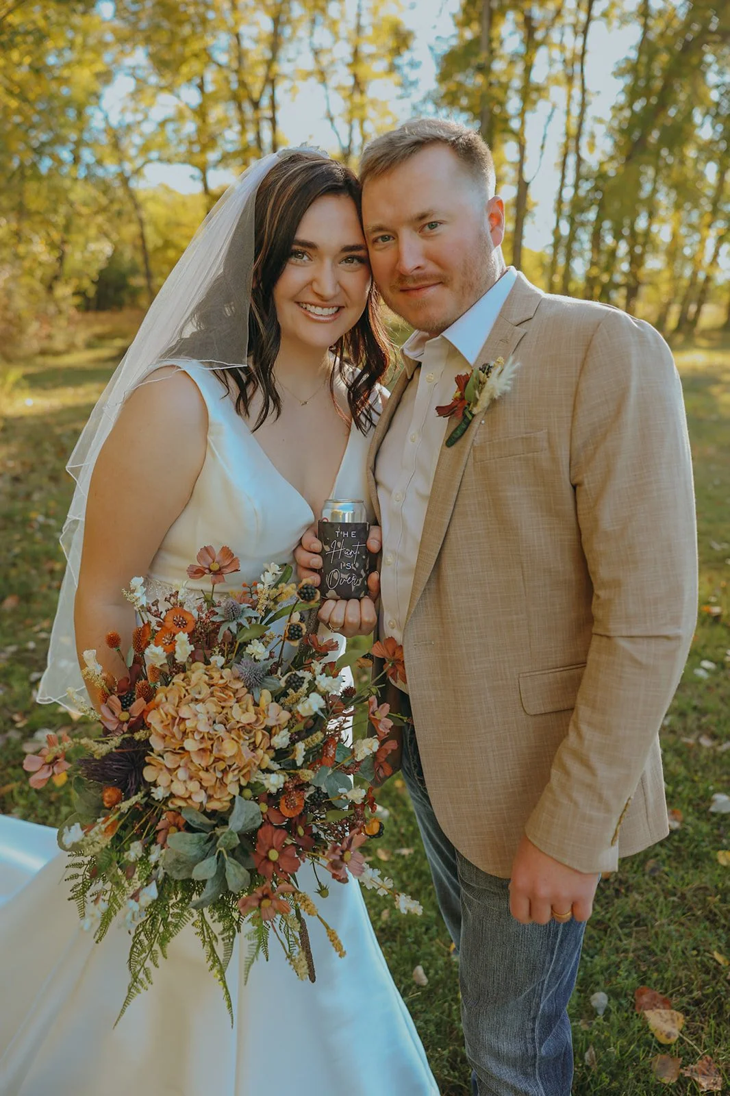 A bride and groom standing outdoors on their wedding day, smiling at the camera. The bride is in a white wedding dress with a veil, holding a large bouquet of fall-colored flowers. The groom is in a beige suit jacket and jeans, holding a drink can wi