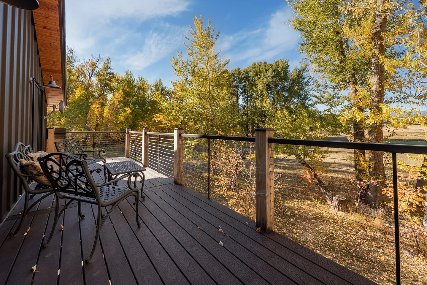 A wooden deck with metal outdoor chairs and a small table, overlooking trees with autumn leaves and a clear blue sky.