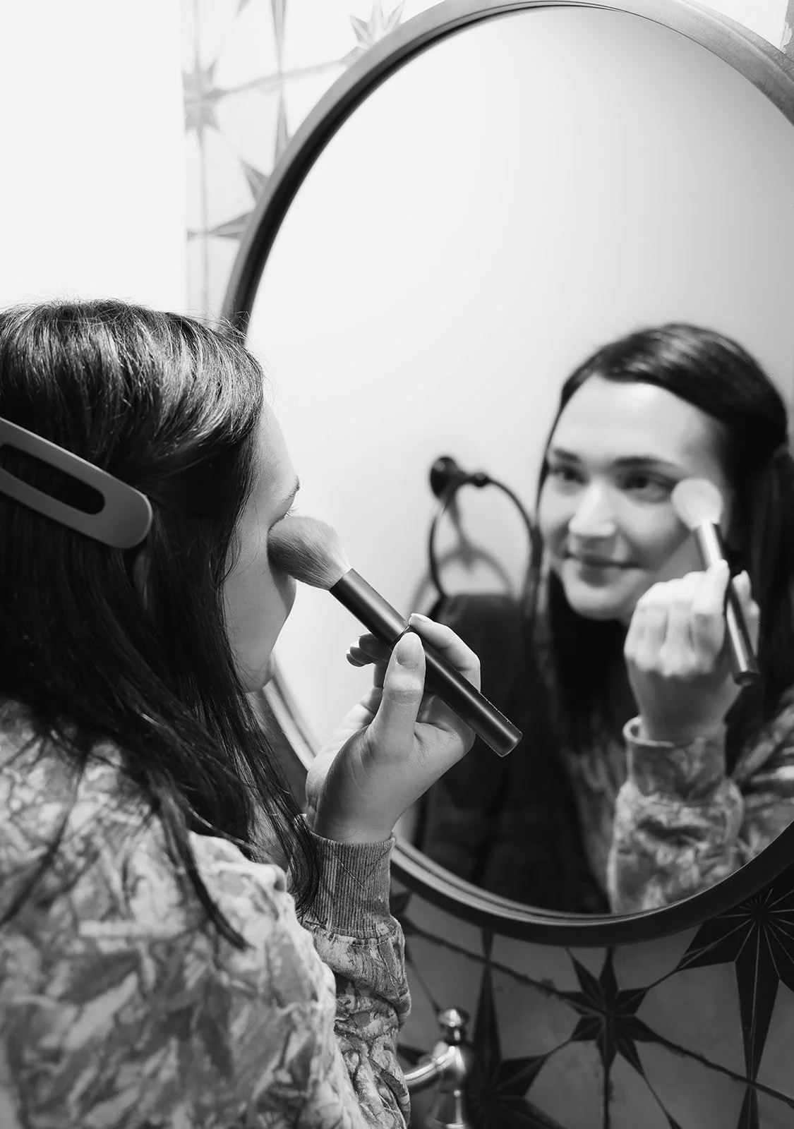 A woman applying makeup using a brush in front of a round mirror.