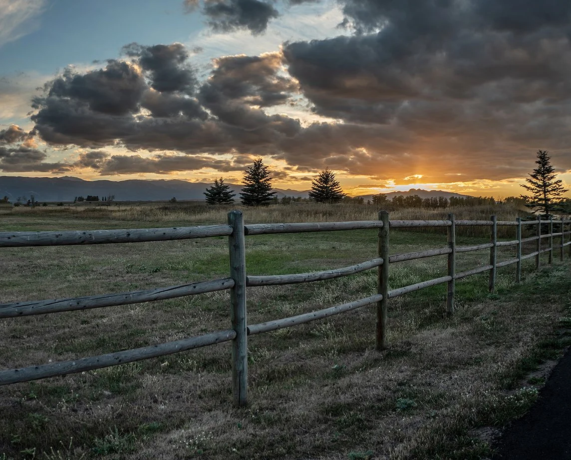 A peaceful rural landscape at sunset with a wooden fence, green grass, trees, mountains in the distance, and a cloudy sky with sunlight breaking through.
