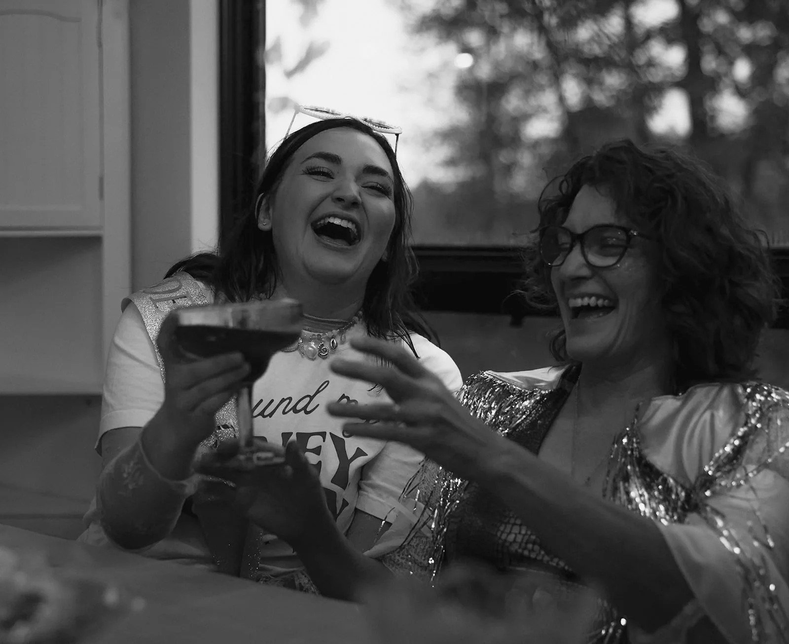 Two women laughing and toasting with drinks at a party, one wearing a sequined outfit and the other wearing a T-shirt with text, in a setting with background window showing trees.