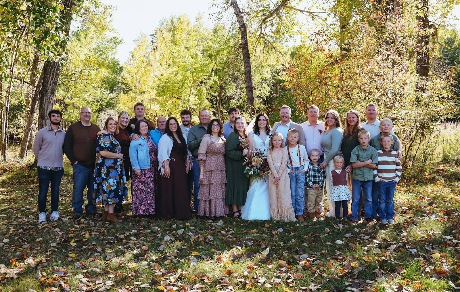 A large group of people outdoors in a wooded area during fall, with trees and fallen leaves; some individuals are dressed for a wedding, possibly a family gathering or celebration.