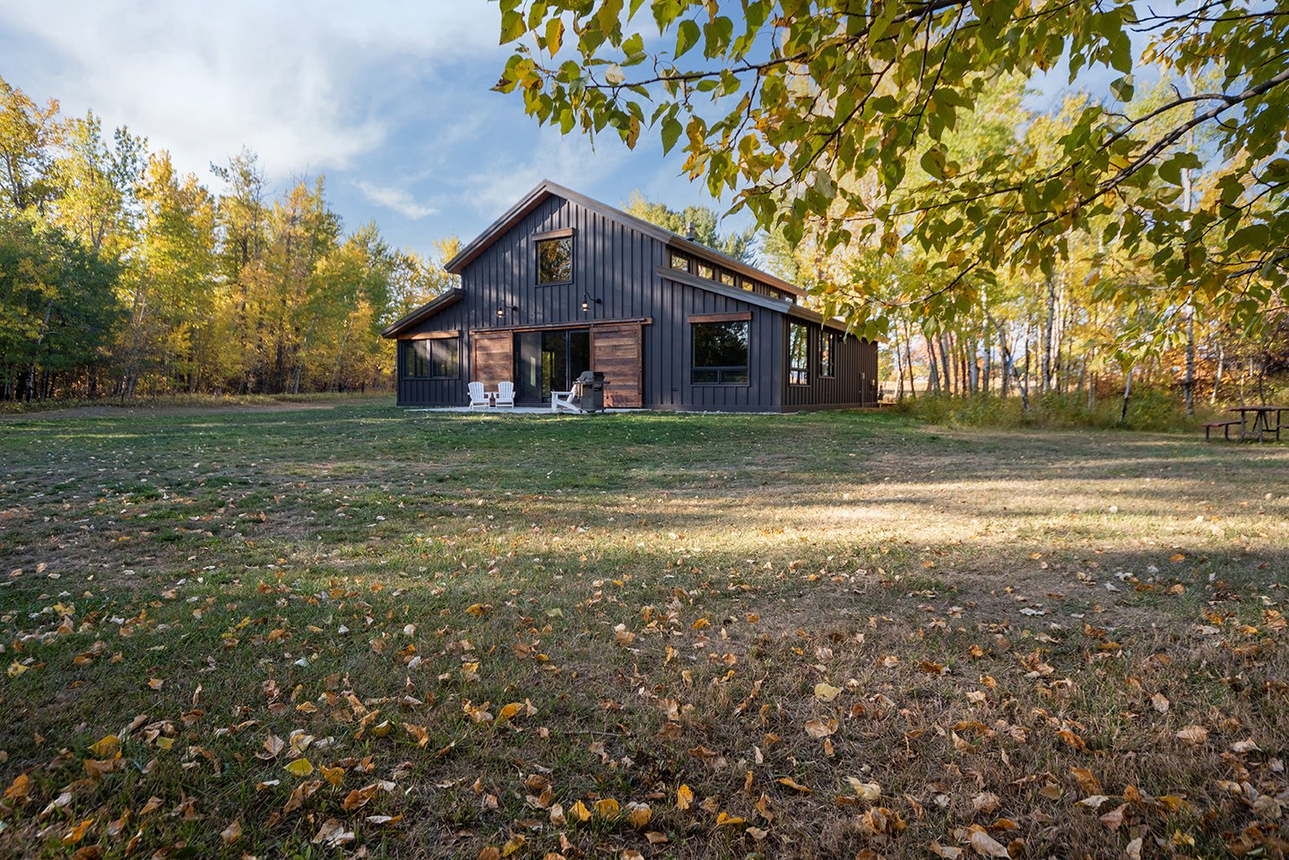 Modern black house with wooden accents, surrounded by trees with autumn foliage, set on a grassy yard with fallen leaves.