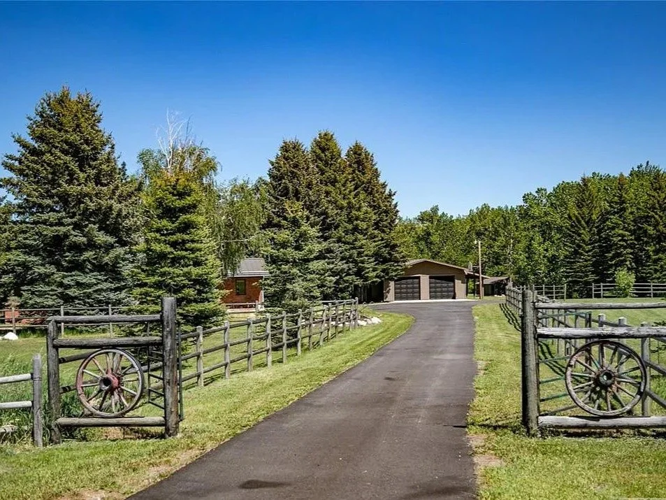 A paved driveway leading to a garage with trees on both sides and a wooden fence with wagon wheels on each side in a rural setting.