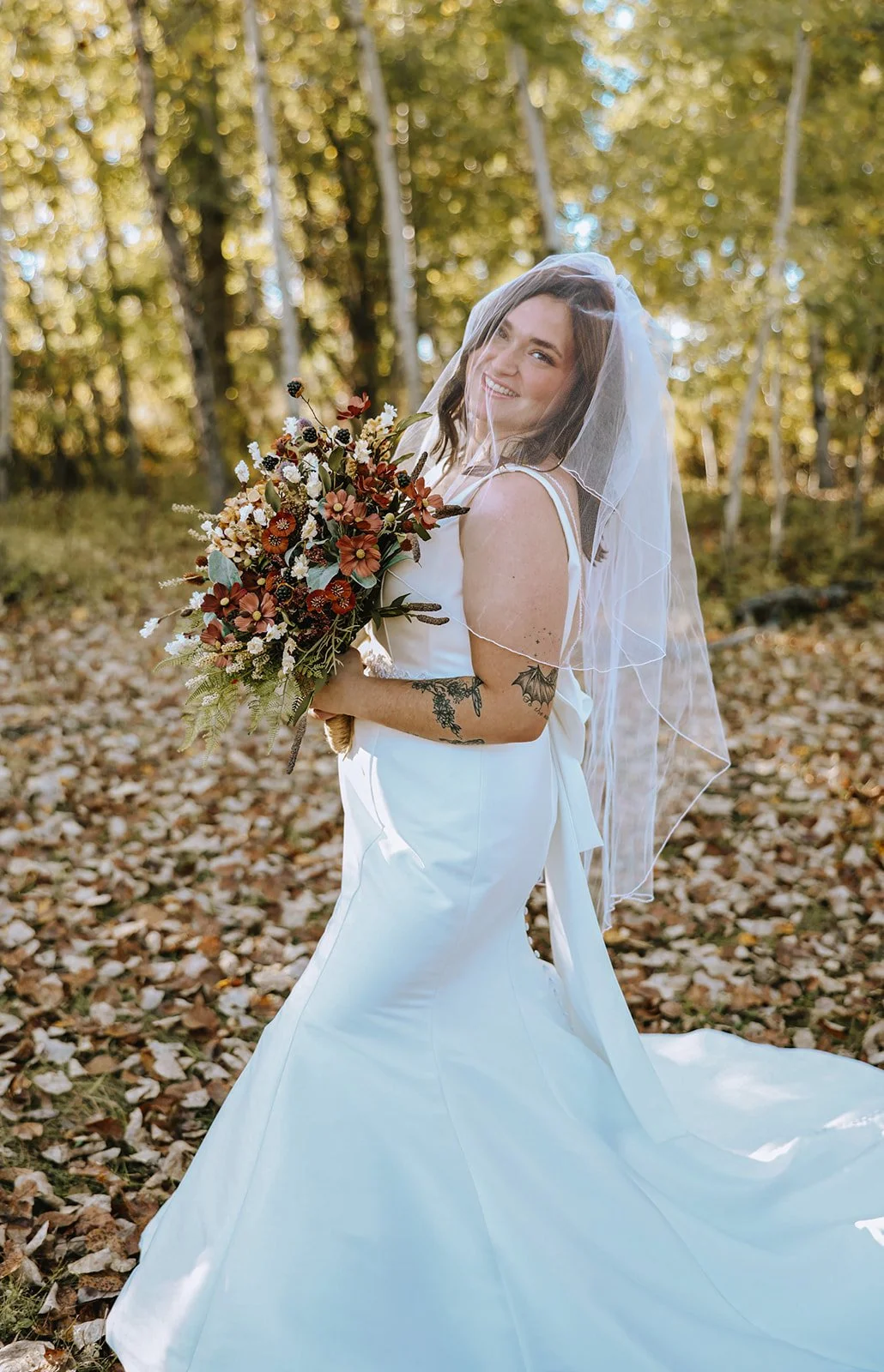 A bride with a veil and tattoos on her arm, holding a bouquet of flowers, standing outdoors among fallen leaves and trees, smiling.