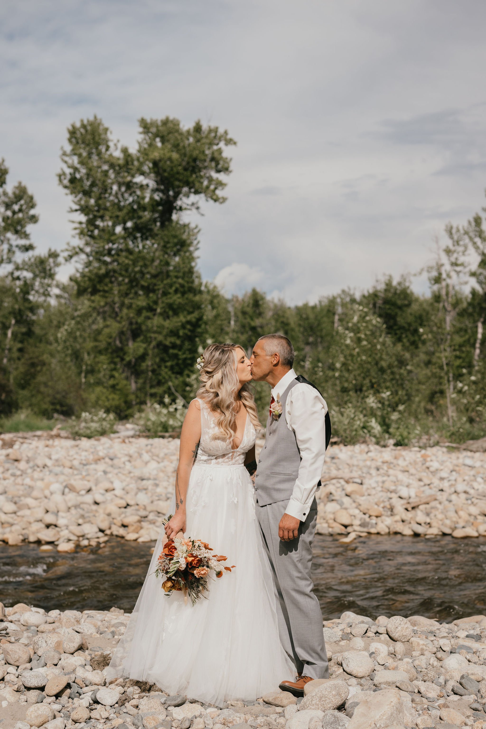 A bride and groom sharing a kiss outdoors by a river with trees and rocks in the background.