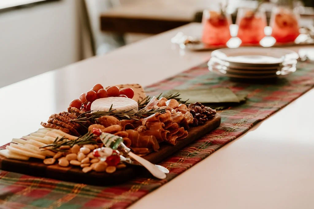 Charcuterie board with cheese, grapes, nuts, and herbs on a table with a holiday-themed table runner.