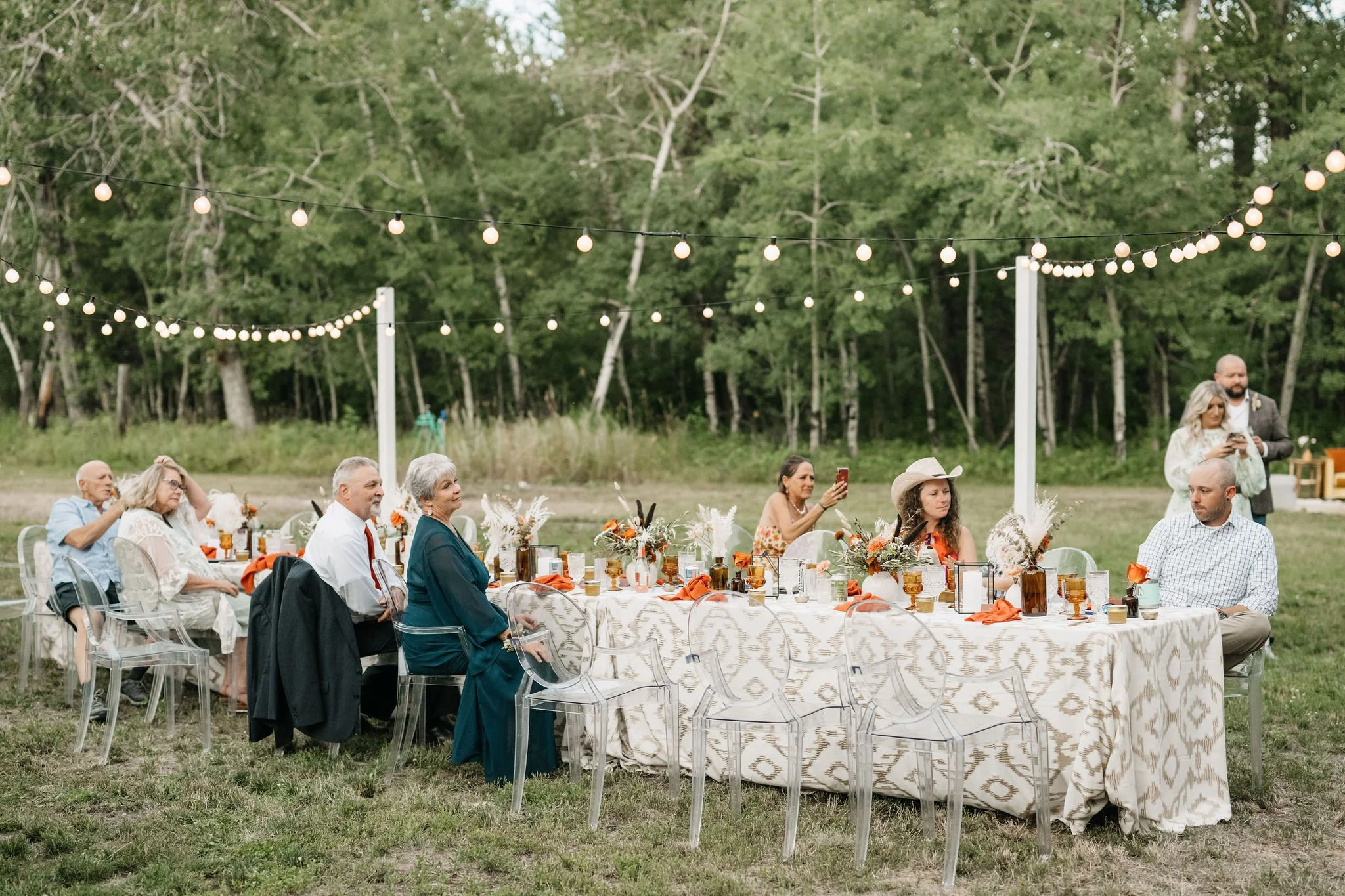 People sitting at a long outdoor wedding reception table decorated with flowers and orange accents, under string lights in a grassy area surrounded by trees.