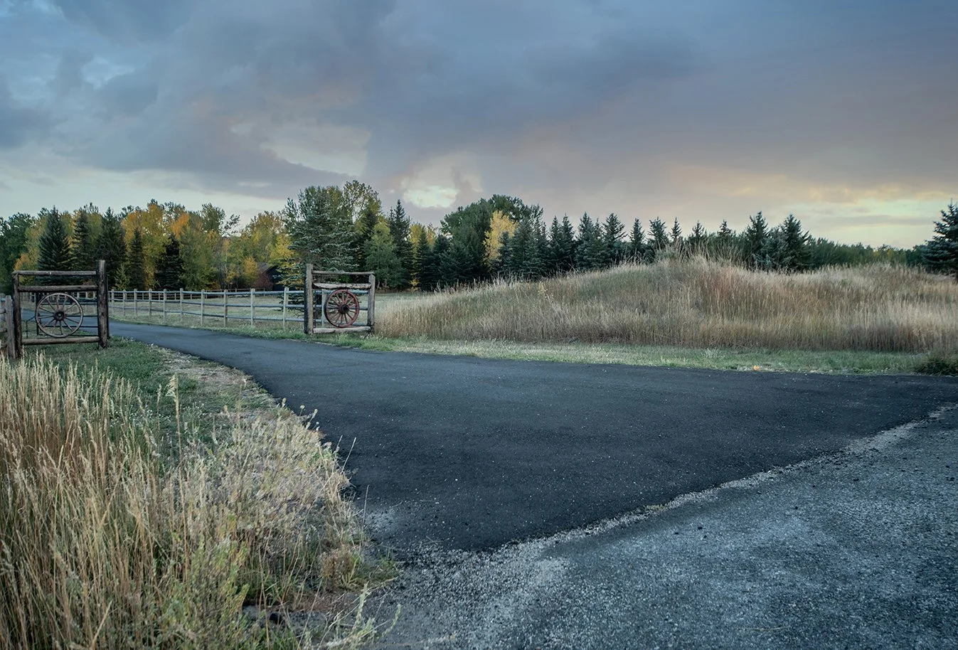 A paved road curves through a rural landscape with tall grass on one side and a wooden fence with wagon wheel accents crossing the road. A line of trees with green and yellow leaves under a cloudy sky is in the background.