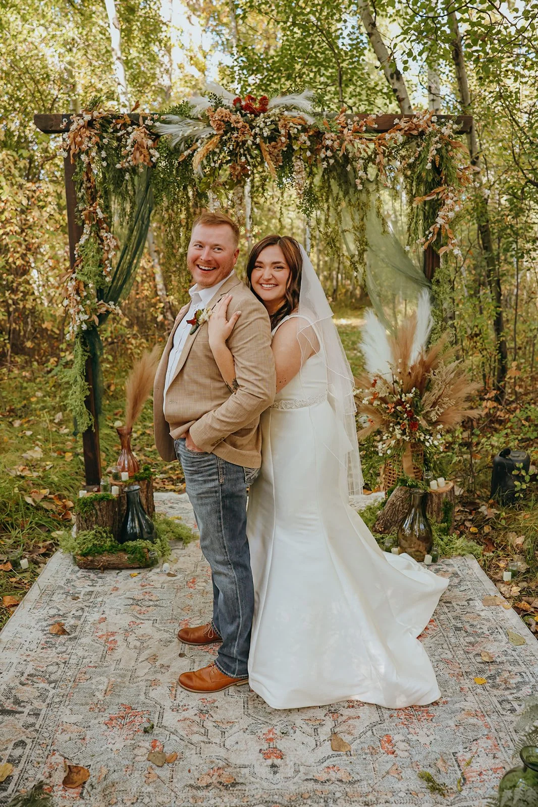 A bride and groom smile and pose for a photo in a wooded outdoor setting during their wedding, standing under a decorated arch with flowers and greenery.