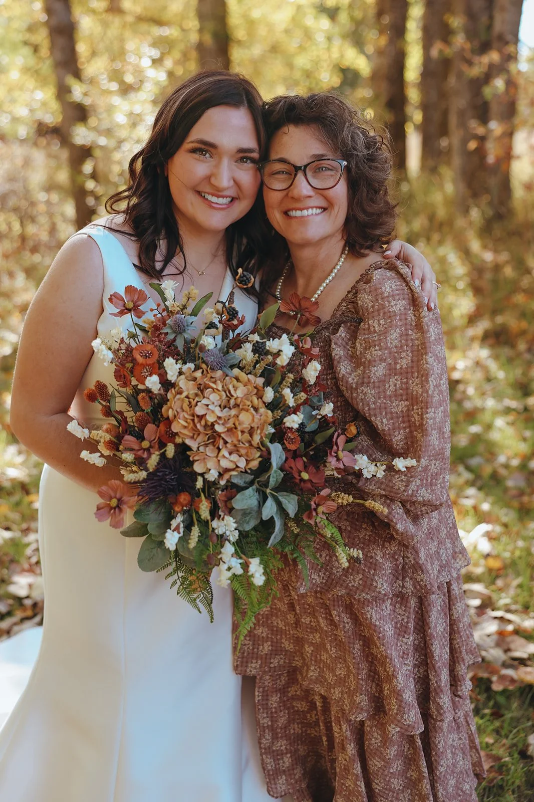 Two women smiling and embracing in a forest; one in a white wedding dress holding a bouquet of flowers, the other in a floral dress with glasses.
