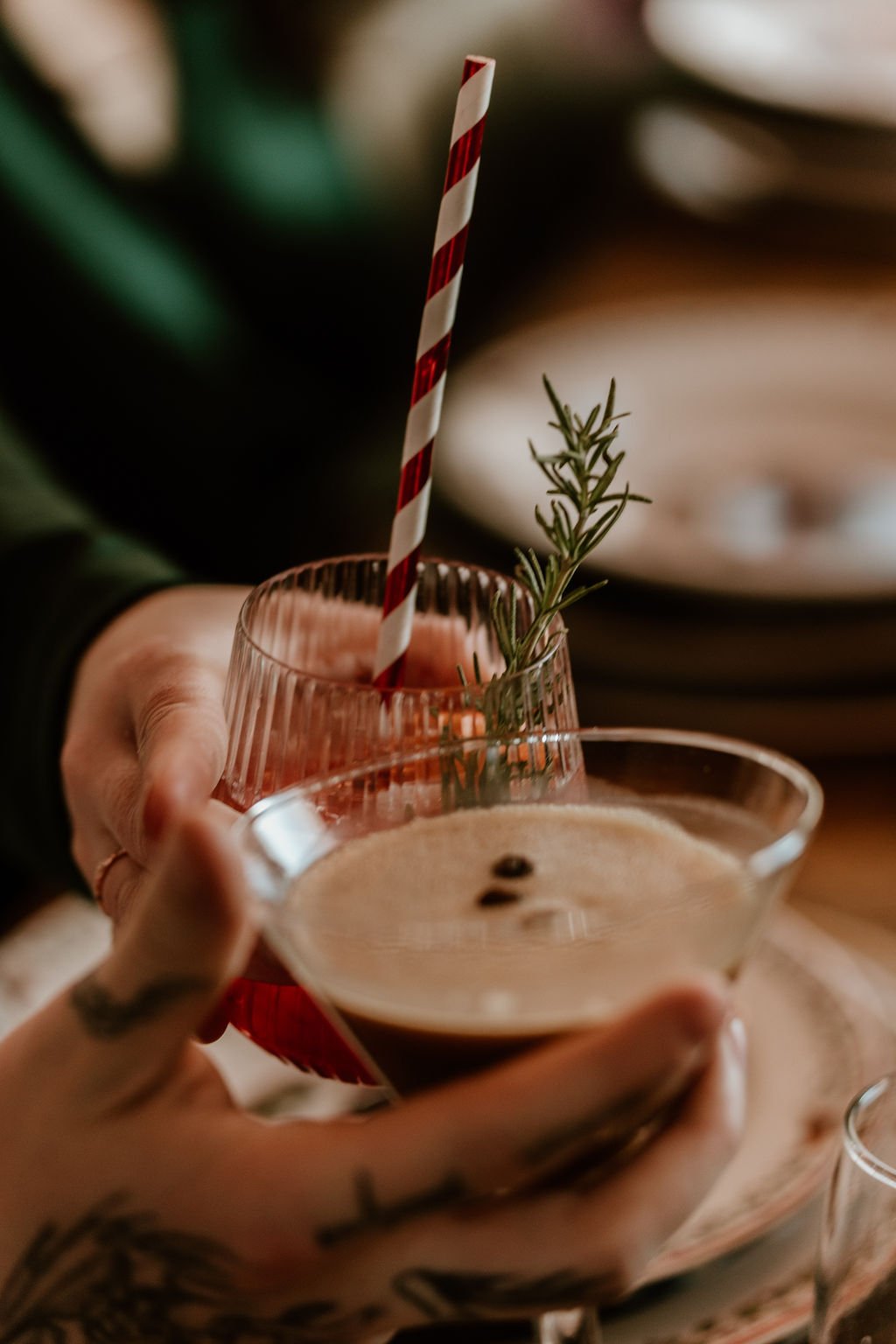 Person holding a glass of coffee with a sprig of rosemary as decoration, and a second glass with a straw and similar garnish in the background.
