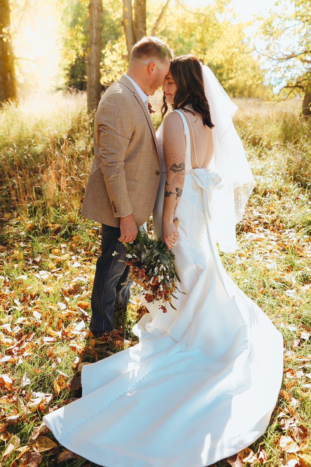 A bride and groom in an outdoor wedding, standing close with foreheads touching, holding a bouquet, surrounded by fall foliage.