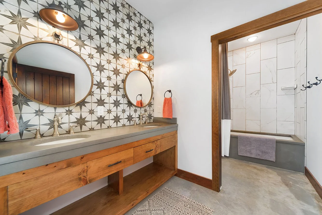 A bathroom with a double vanity featuring a concrete countertop, two oval mirrors, and wall-mounted lights. The wall behind the vanity has a geometric tile pattern. To the right, there is a shower area with white large tiles, a built-in niche, and a 
