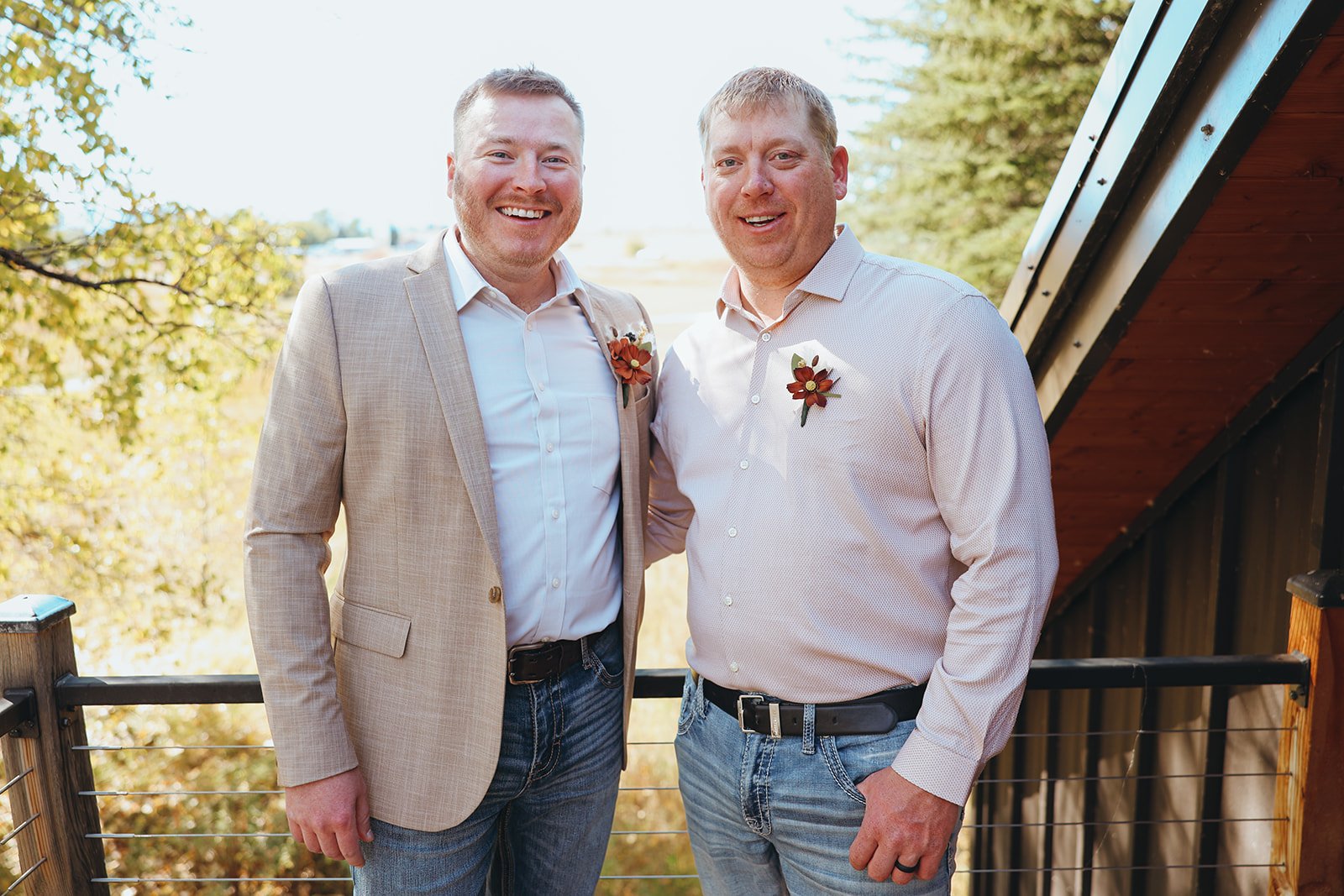 Two men dressed in light-colored shirts and jeans, standing outdoors on a wooden balcony, smiling. They both have boutonnières on their shirts, indicating a special occasion such as a wedding. The background features autumn trees and a clear sky.
