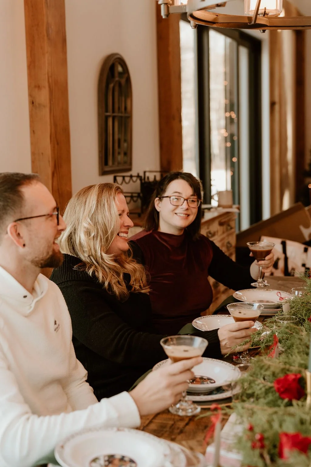 Three people sitting at a decorated table during a holiday gathering, holding drinks, smiling, in a cozy, warmly lit room.