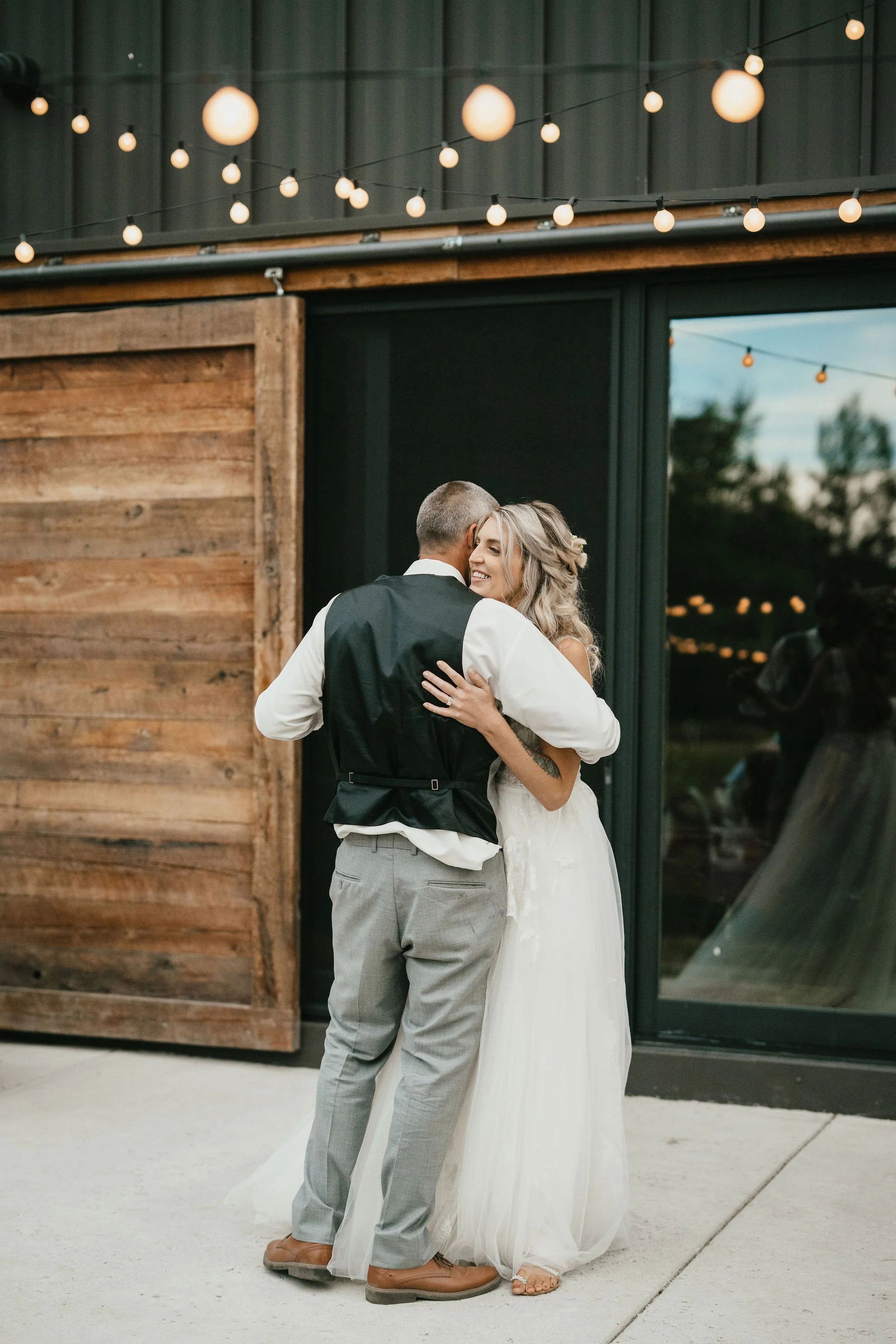 A bride and groom hugging during their wedding reception outside. The bride is smiling and wearing a white wedding dress, while the groom is wearing a black vest and gray pants. String lights are hanging above them against a backdrop of a barn with s