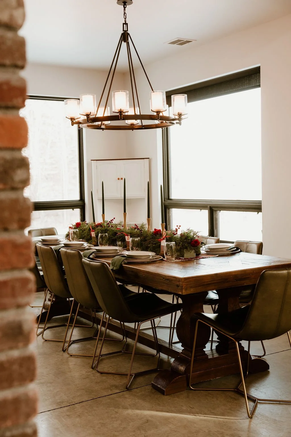 A dining room with a wooden table decorated with holiday greenery and candles, surrounded by black chairs, and lit by a chandelier, with large windows bringing in natural light.