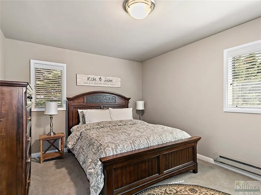 A bedroom with a wooden bed frame, white bedding, two nightstands with lamps, and two windows with white blinds, beige walls, and a ceiling light.