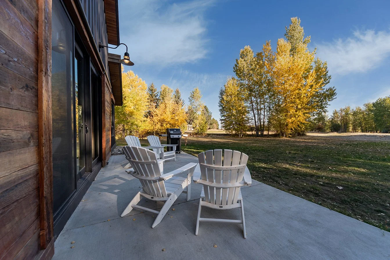 An outdoor patio with four white Adirondack chairs, a barbecue grill, and a wooden house with sliding glass doors, surrounded by trees with yellow and green leaves under a blue sky.