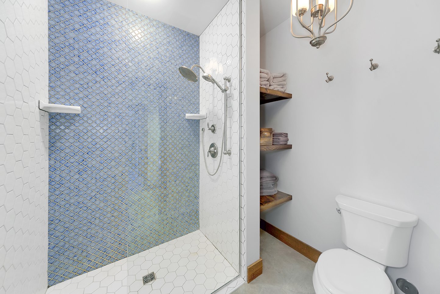 Bathroom with a walk-in shower featuring a blue mosaic tiled wall. The shower has a rain shower head and two small white shelves. Adjacent to the shower is a white toilet and wooden shelves with folded towels. The bathroom has white hexagonal floor t