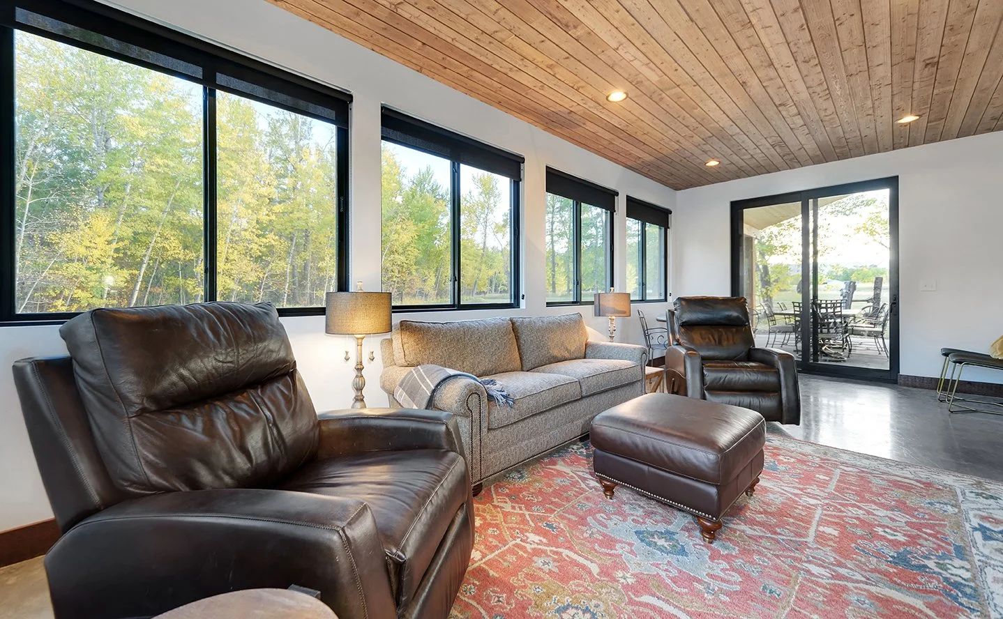 Living room with leather recliners, a gray sofa, lamps, large windows, and a sliding glass door leading to an outdoor patio with a dining table.