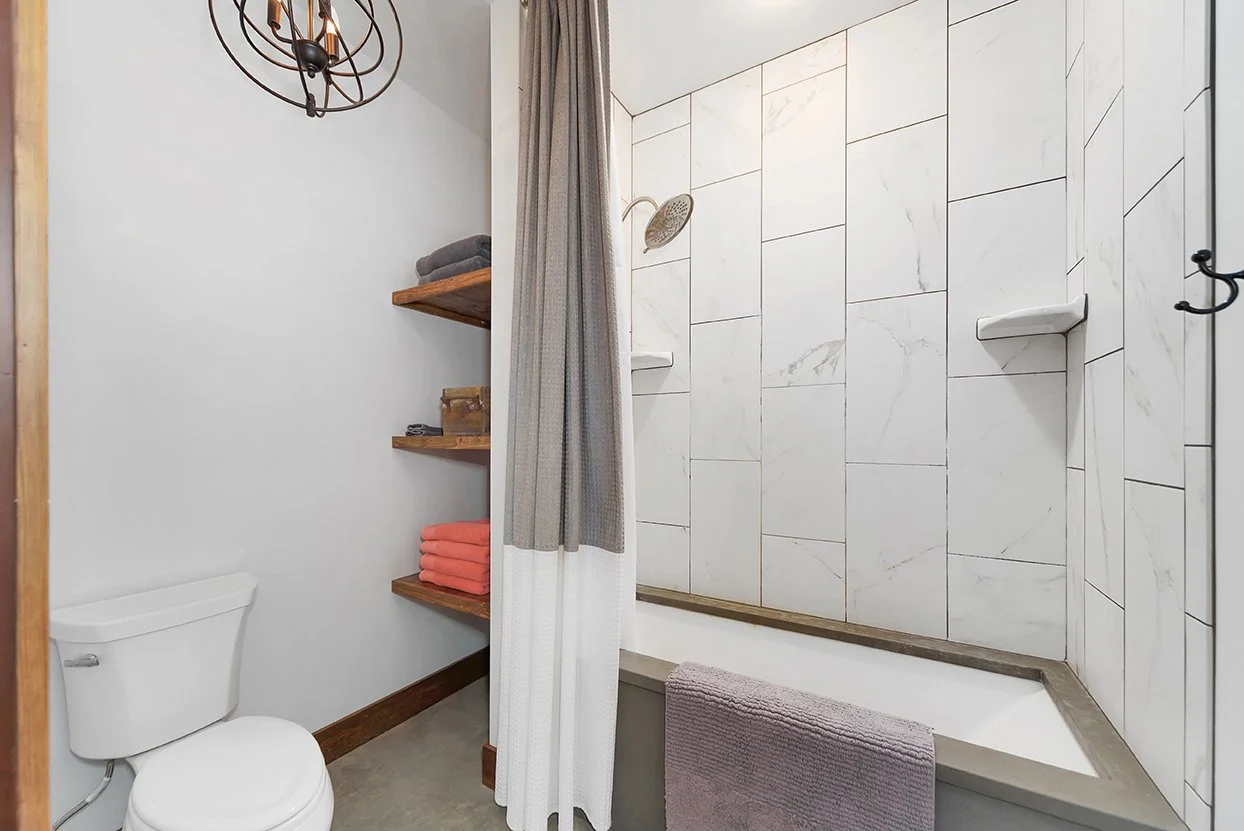 A modern bathroom featuring a shower with white marble tiles, a built-in shelf, a purple towel hanging on the edge of the tub, a toilet, a wooden shelf with towels and toiletries, and a stylish metal light fixture.