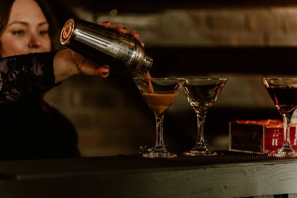 A bartender pours a cocktail from a shaker into a martini glass, with two other cocktail glasses nearby on a bar counter.