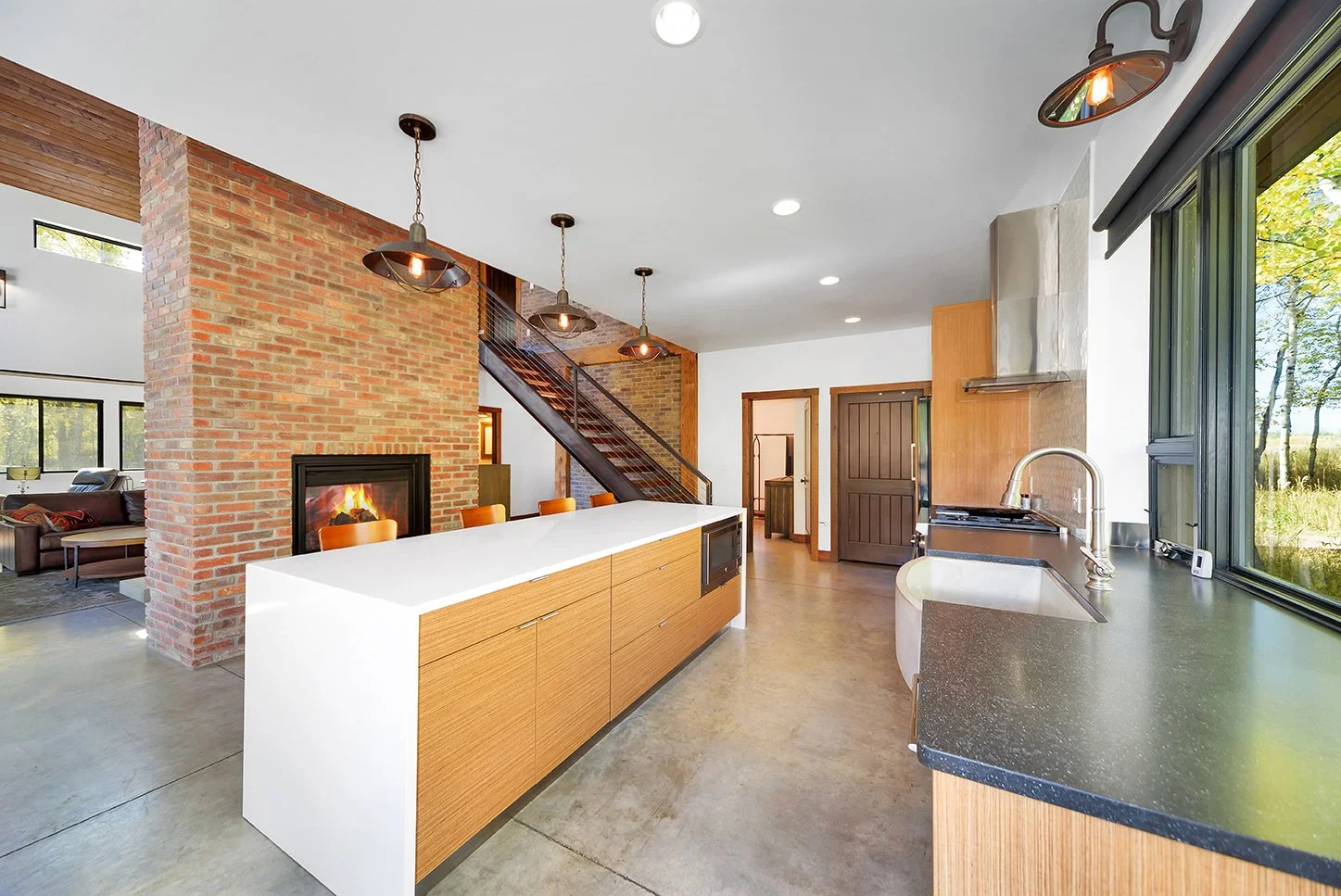 Modern kitchen with brick wall, white and wood cabinets, large window showing outdoor scenery, black countertop, and stainless steel appliances, with pendant and recessed lighting.
