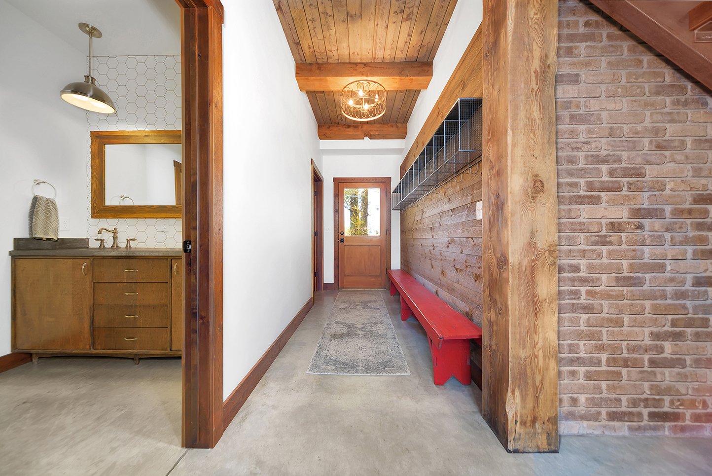 Entryway with a brick wall on the right, a wooden ceiling, a red bench alongside the wall, and a door at the end. To the left, part of a bathroom with a wooden vanity, a mirror, and a hexagonal tile wall, visible through a doorway.