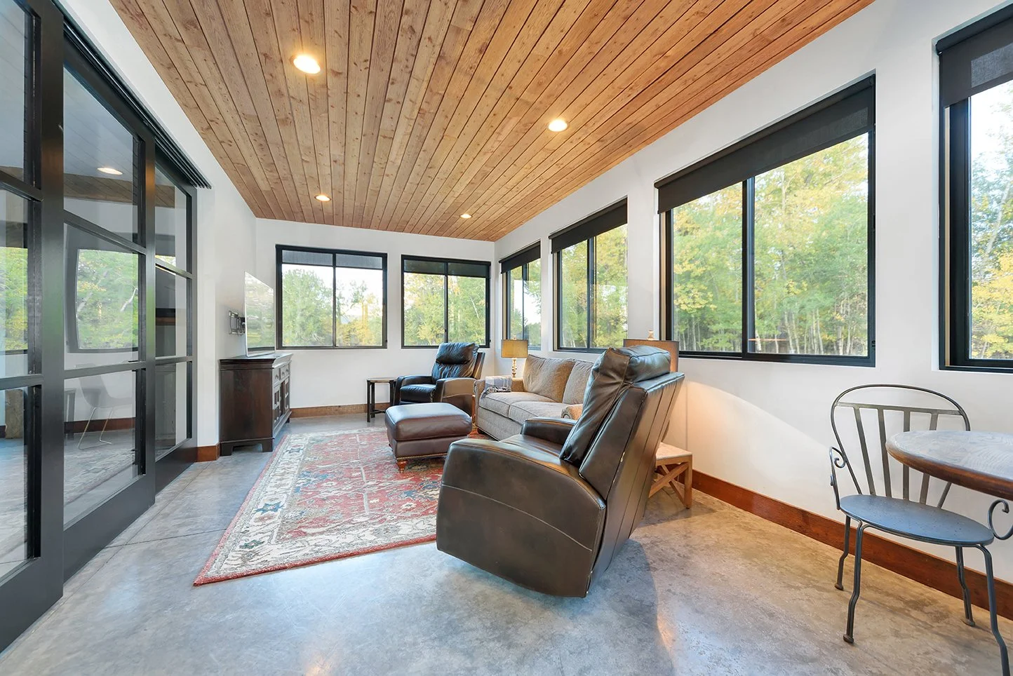 Sunroom with leather armchairs, a sofa, a patterned rug, and large windows with black frames showing trees outside.