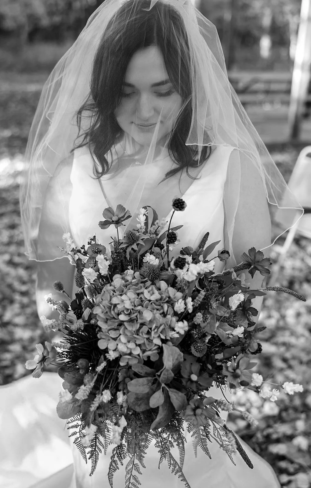 Black and white photo of a bride holding a bouquet of flowers, wearing a wedding dress and veil, outdoors.